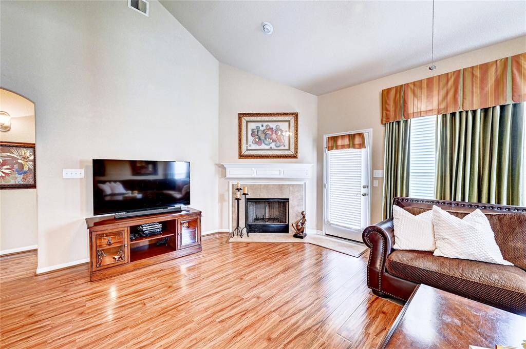 This cozy living room features a high ceiling, hardwood floors, and a welcoming fireplace. It includes a large window with elegant drapes, a comfortable sofa, and a TV setup. Ideal for relaxation and entertaining.
