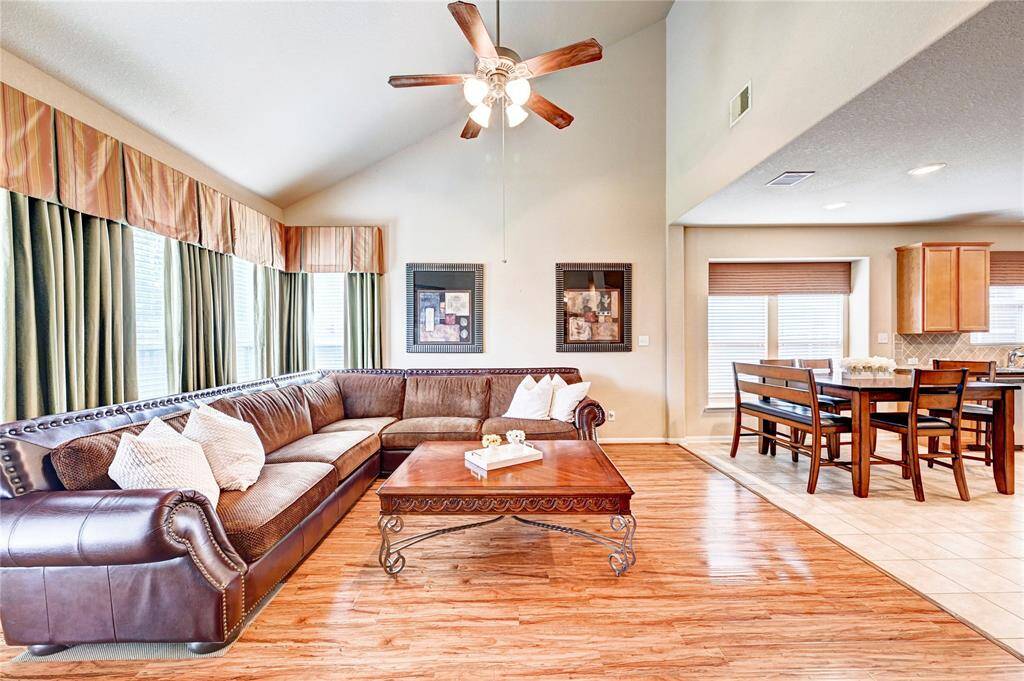 This spacious living area features a high ceiling with a ceiling fan, enough room for a  large sectional sofa, and has abundant natural light from multiple windows. It seamlessly flows into an open dining area with a wooden table and chairs, creating a warm and inviting space perfect for entertaining.