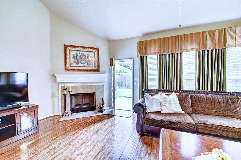 Bright living room with hardwood floors, a cozy fireplace, and large windows with drapes. Features a comfortable sofa, a TV on a wooden stand, and access to a backyard through a glass door.