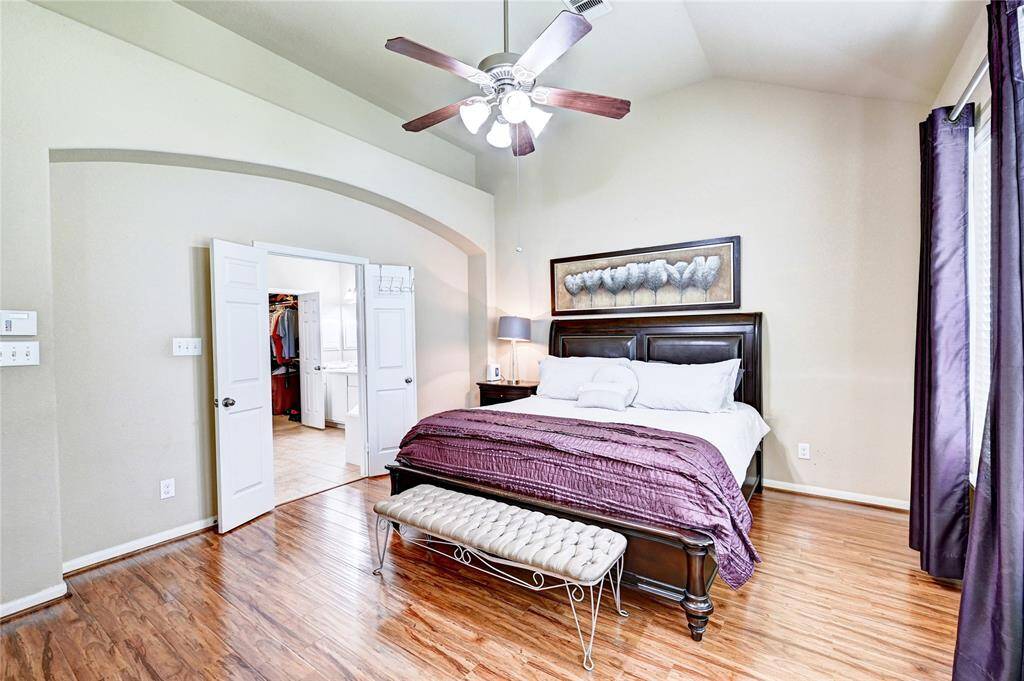 This photo shows a spacious bedroom with a high ceiling and hardwood floors. The room has ample natural light from large windows. There's an entrance to the  bathroom, enhancing convenience and privacy. A ceiling fan adds comfort, and tasteful decor completes the inviting space.