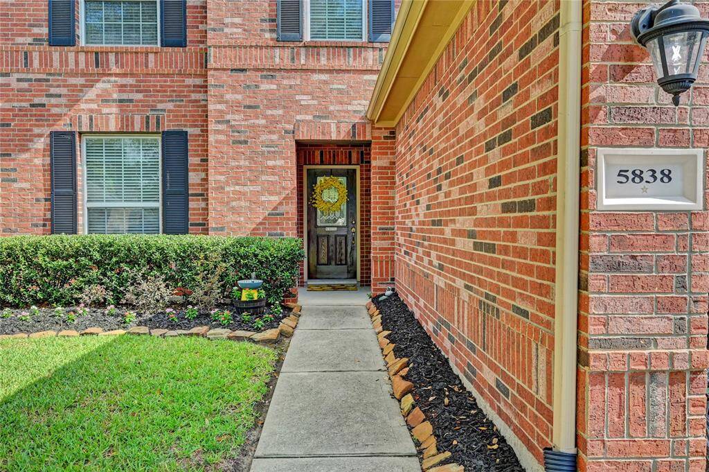 This photo showcases the welcoming front entrance of a brick home, featuring a well-maintained walkway lined with trimmed hedges and a manicured lawn. The charming front door has a decorative glass.