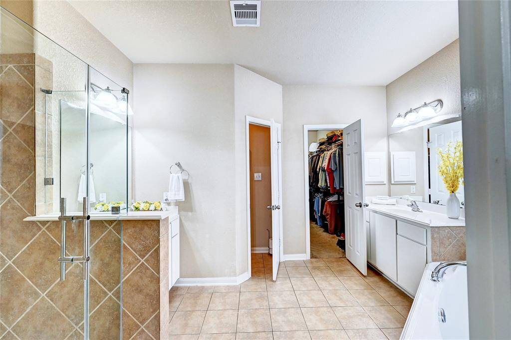 This spacious bathroom features a walk-in shower with glass doors, dual sinks with ample counter space, and a large walk-in closet. The neutral tiles and soft lighting create a warm and inviting atmosphere.
