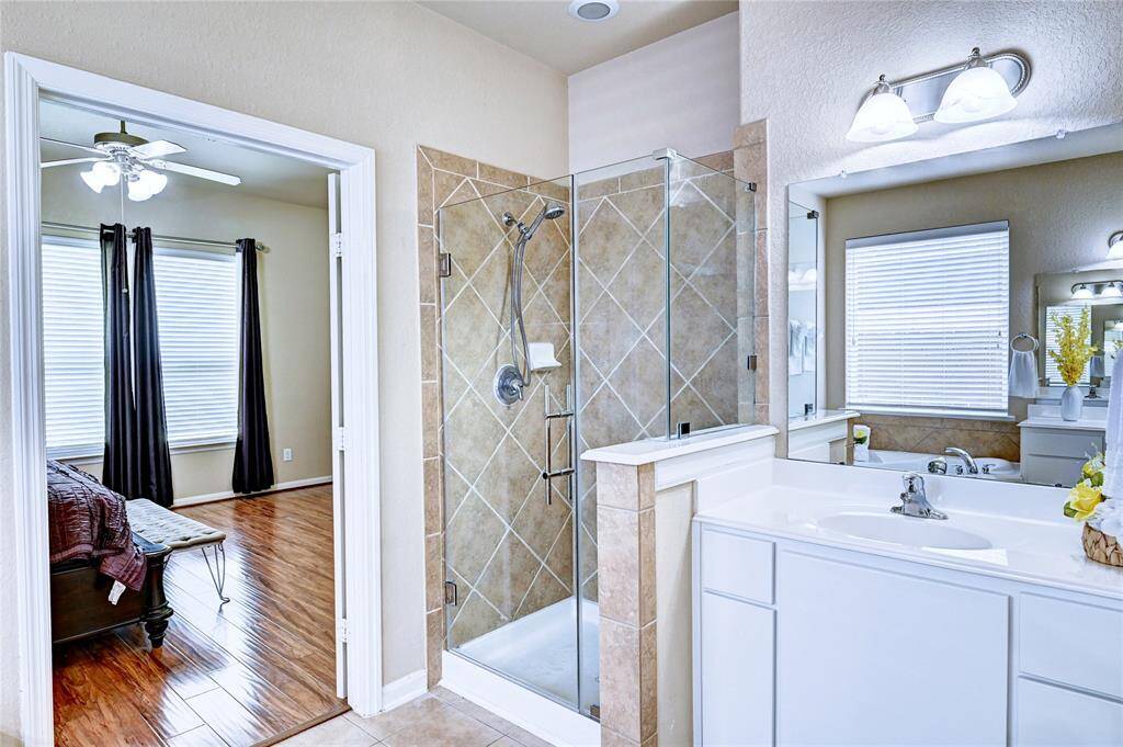 This photo showcases a bright and modern bathroom adjacent to a bedroom. The bathroom features a glass-enclosed shower with tile accents, a sleek white vanity with ample storage, and overhead lighting. The adjacent bedroom has wooden flooring, large windows with blinds,  creating a cozy yet elegant atmosphere.
