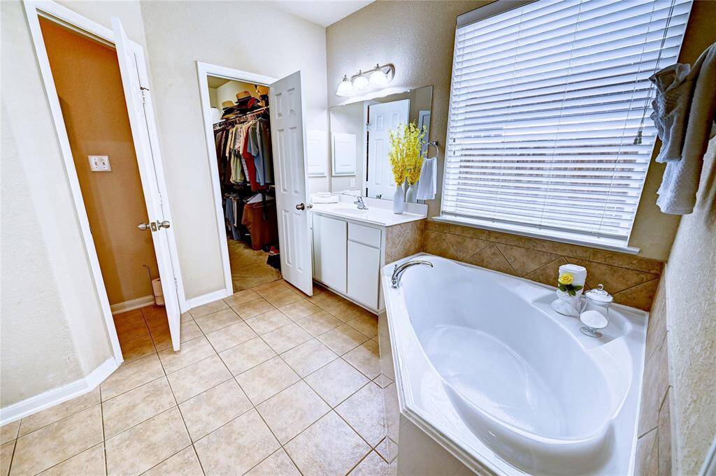 This bathroom features a corner soaking tub with elegant tile surround, a single vanity with a large mirror, and ample natural light from the window. It connects to a spacious walk-in closet, providing convenience and functionality.