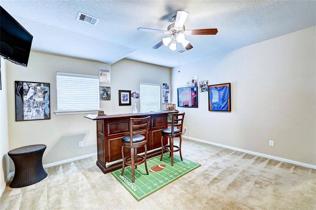 This photo showcases a cozy entertainment room featuring a small bar with two stools, sports-themed decor, and carpeted flooring. The room is well-lit with a ceiling fan and has two windows. Ideal for hosting gatherings or relaxing.