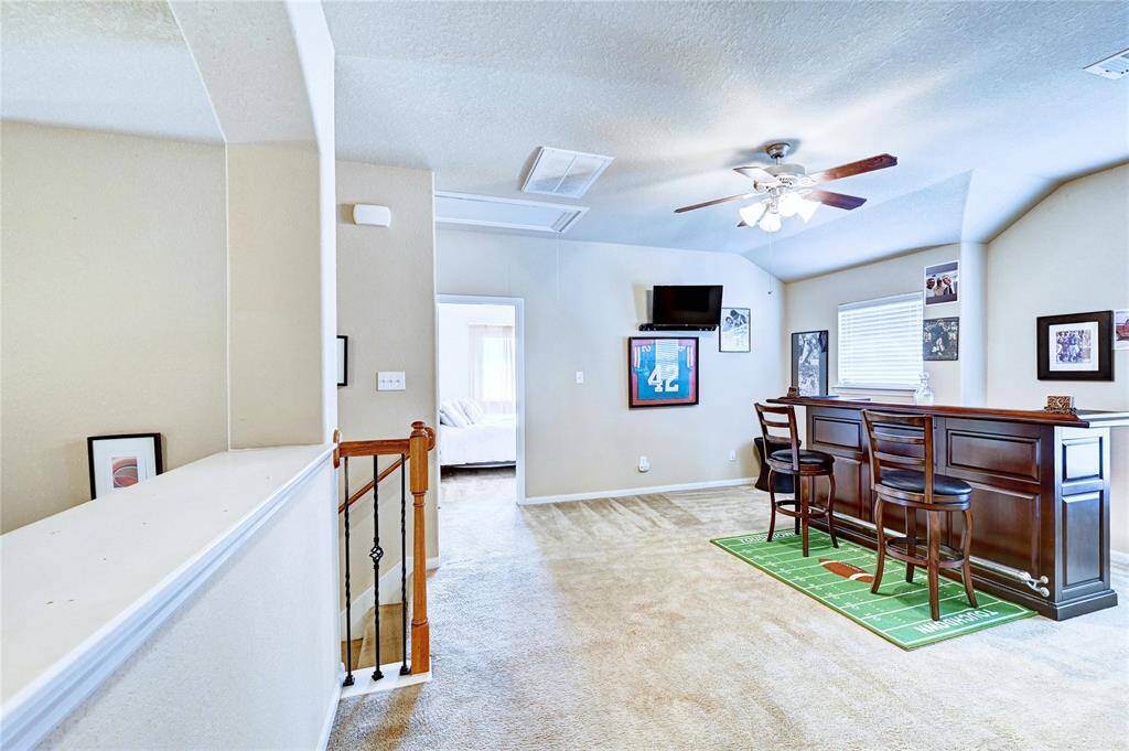 This room features a cozy entertainment area with a bar setup and high stools. It has a ceiling fan, carpeted floors, and sports-themed decor, offering a casual and inviting space.