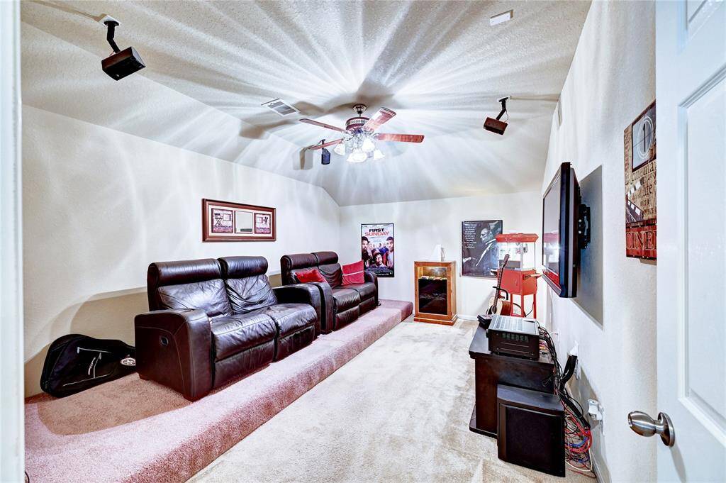 This photo shows a cozy home theater room featuring comfortable black leather recliners, a large wall-mounted TV, surround sound speakers, and movie-themed decor. The room has carpeted floors and a ceiling fan, creating an inviting atmosphere for entertainment.