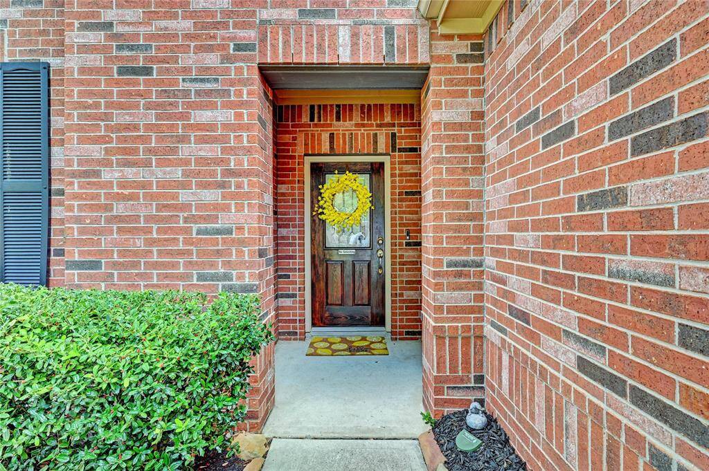 Charming brick entryway with a welcoming wooden door adorned with a bright wreath. The area features a tidy hedge and a cozy porch with a decorative mat, offering a warm and inviting first impression.