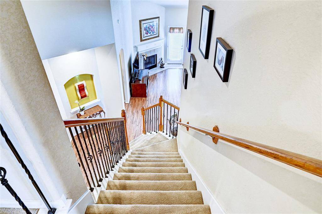 From the game room, this photo shows a view from the top of a carpeted staircase with wooden handrails leading down to a bright, open living area with hardwood floors. Kitchen area is to the right at the bottom of the stairs.