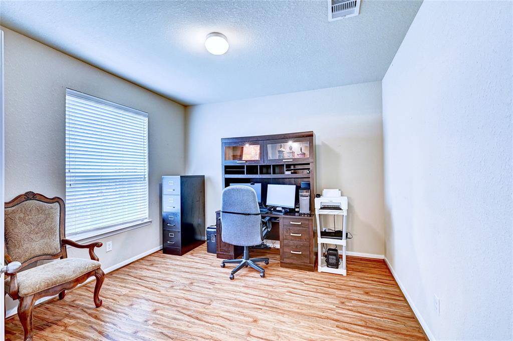 A bright home office with wood flooring. Natural light streams in from a large window with blinds.