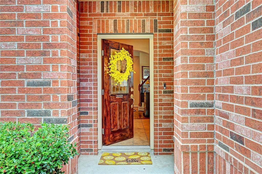 Charming brick entryway with a welcoming wooden door adorned with a bright yellow wreath. A small doormat adds a cozy touch, and the interior glimpse shows a staircase, enhancing the home's inviting atmosphere.