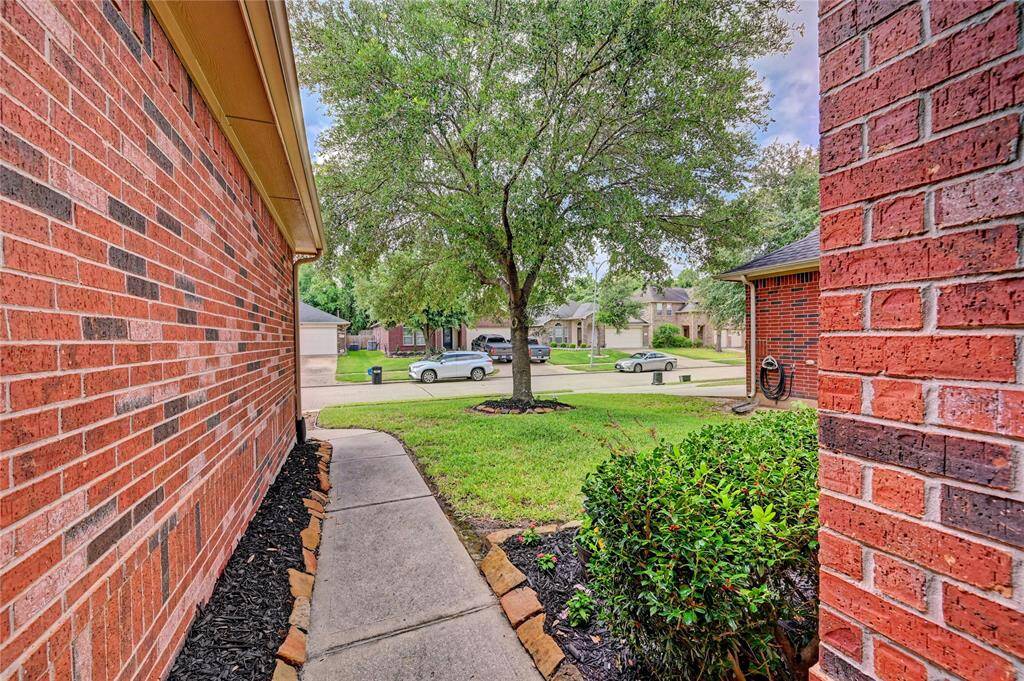 This photo shows a charming front yard with a paved walkway bordered by red brick walls. There's a neat lawn with a mature tree, and the view opens up to a quiet suburban street with neighboring houses.