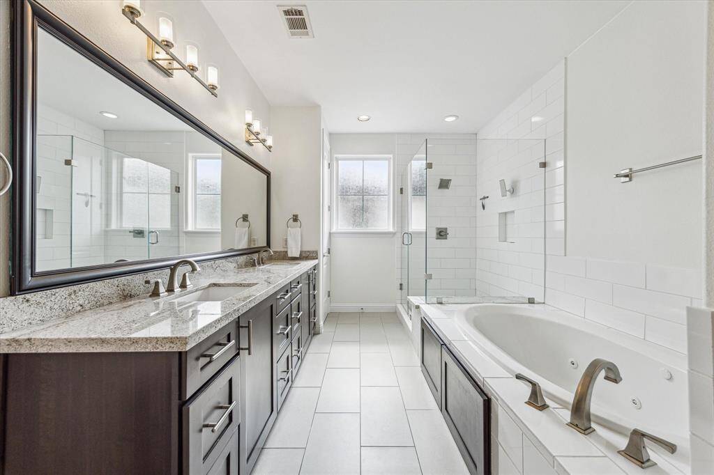 The spa-inspired ensuite offers a soothing escape with a deep jetted tub, an oversized frameless glass shower, and a dual-sink granite vanity topped with a framed mirror and designer lighting.