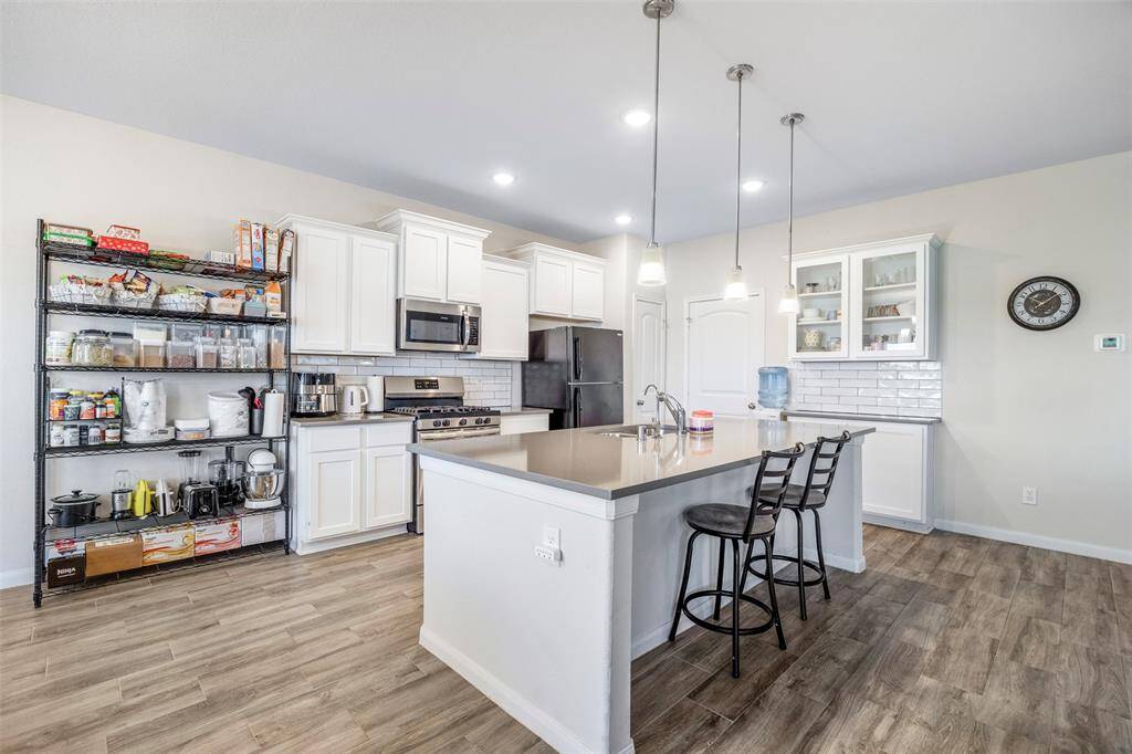 This modern kitchen features white cabinetry, stainless steel appliances, and an island with pendant lighting and bar seating. The space is enhanced by a sleek backsplash, hardwood-style flooring, and a cozy, open layout.