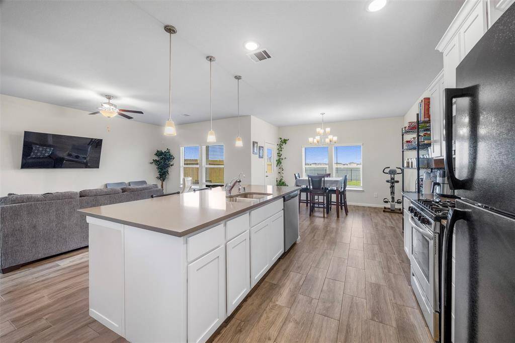 This photo showcases an open-concept kitchen and living area with modern features. The kitchen has a large island with pendant lighting, white cabinetry, and stainless steel appliances. The space flows into a cozy living room with a ceiling fan and overlooks the breakfast area. Large windows provide ample natural light, and wood-look flooring adds warmth throughout.