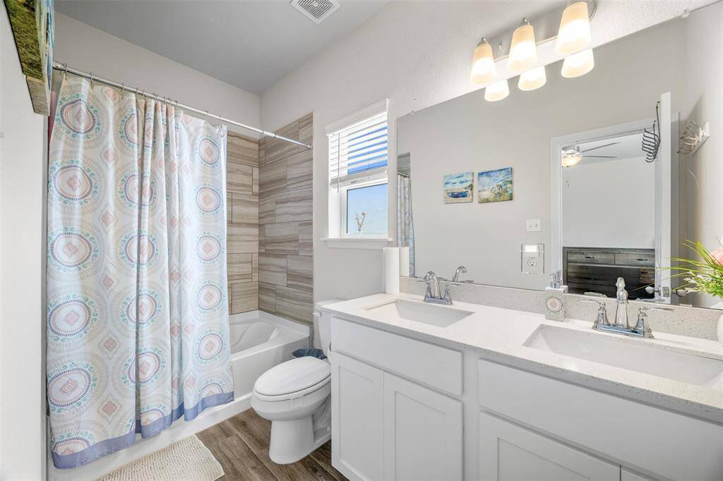 This bathroom features a clean and modern design with a double vanity, ample counter space, and a large mirror. It includes a bathtub with a stylish tiled surround. The room is well-lit, with a window allowing natural light, and has a light wood floor that complements the neutral walls.