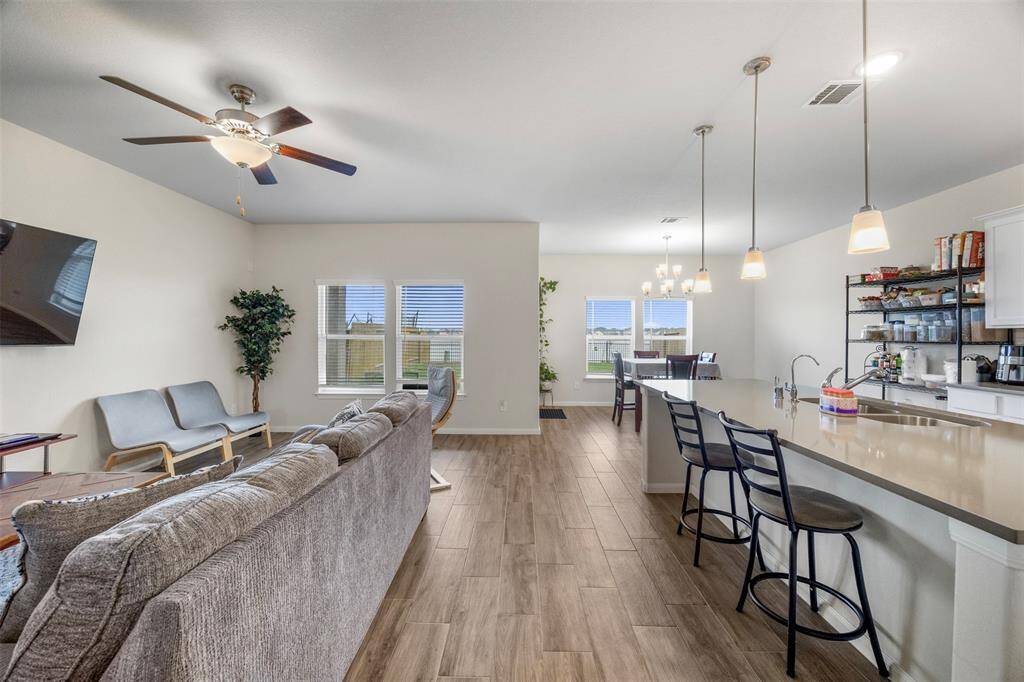 This open-concept living space features a cozy seating area with a ceiling fan, a modern kitchen with a large island and bar seating, and a dining area with natural light from multiple windows. The wood-look tile flooring adds warmth and continuity throughout the space.