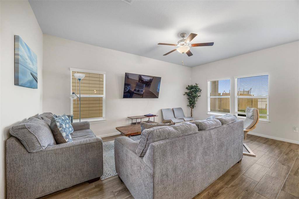 This inviting living room features an abundance of wall space for your TV or Art, and large windows for natural light. The space is complemented by a ceiling fan, wooden flooring, and a cozy seating arrangement idyllic for relaxation and entertainment.