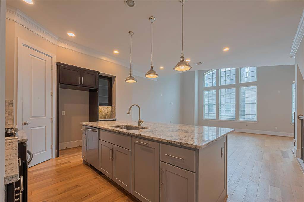 View from the kitchen into the open living area, showcasing a large granite island with undermount sink, stylish pendant lighting, and ample cabinetry. The soaring ceilings and wall of windows in the adjacent living space flood the home with natural light, creating an inviting and airy atmosphere.