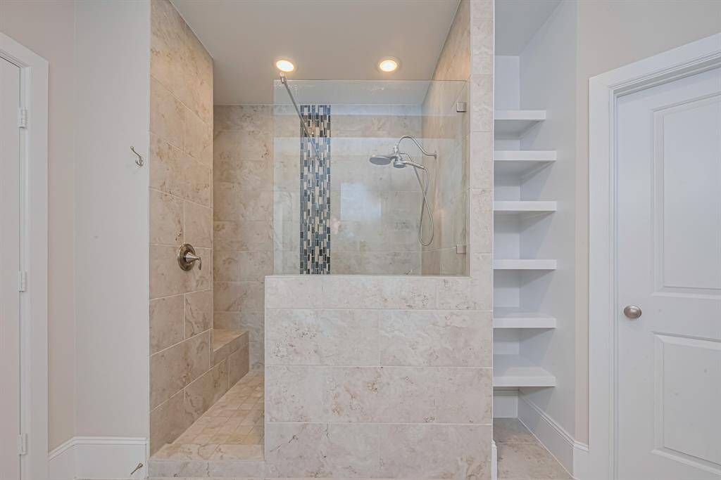 The primary bathroom features a spacious, walk-in shower with custom tile accents, a built-in bench, and dual shower heads. Just steps away, a long granite double-sink vanity provides plenty of storage and prep space. A large walk-in closet with built-in shelving is also accessible directly from the bathroom, offering excellent functionality and organization.