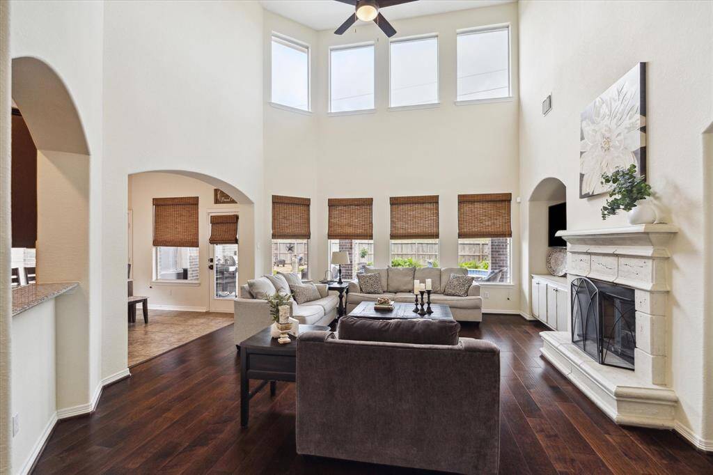 This stunning living room features towering high ceilings, offering a sense of grandeur and openness. A generous wall of windows with woven shades bathes the room in natural light, accentuating the sleek, dark hardwood flooring. The space includes a beautifully crafted stone fireplace and an open connection to the kitchen and dining areas, perfect for modern living and entertaining.