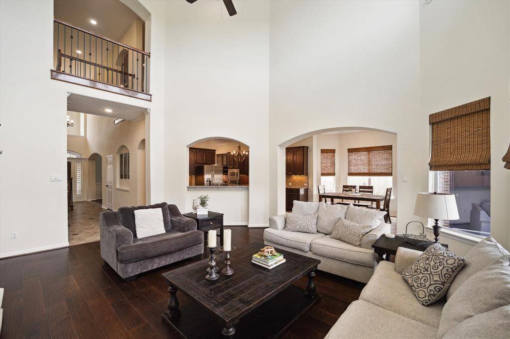 This inviting living room showcases soaring high ceilings that create a grand, open feel. A striking wall of windows floods the space with natural light, highlighting the rich, dark hardwood flooring with an upper-level balcony overlooking the area, enhancing its spacious appeal.