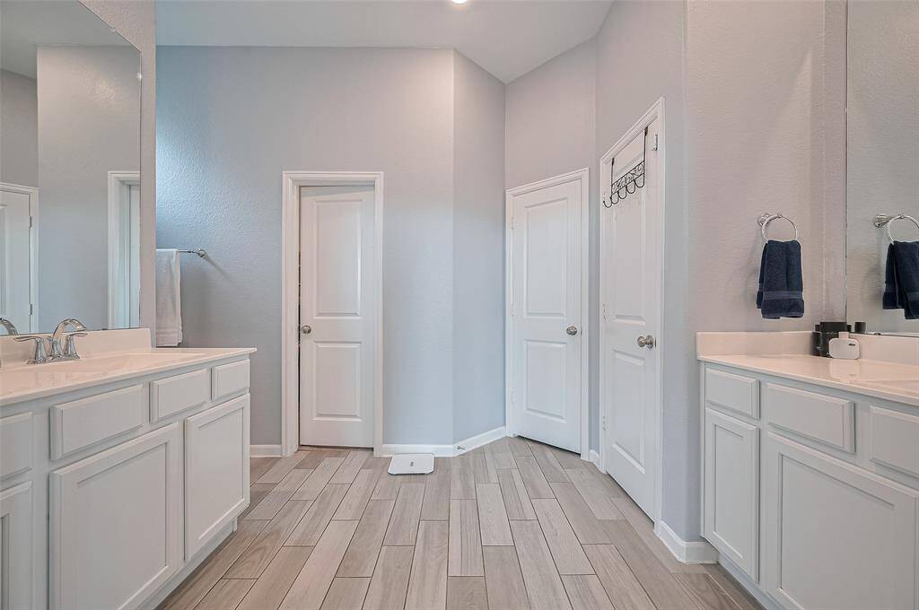 The spacious primary bath showcases dual vanities, ample cabinetry, and wood-look tile floors, offering both function and elegance. Crisp white finishes and soft lighting add to the serene, spa-like atmosphere