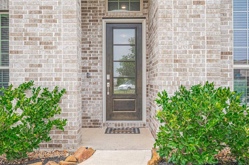 Inviting 8 foot front entry showcases a sleek, glass-paneled door with a transom window above, allowing soft natural light to brighten the space. Framed by rich brick detailing and lush, manicured greenery, it offers a polished and welcoming first impression.