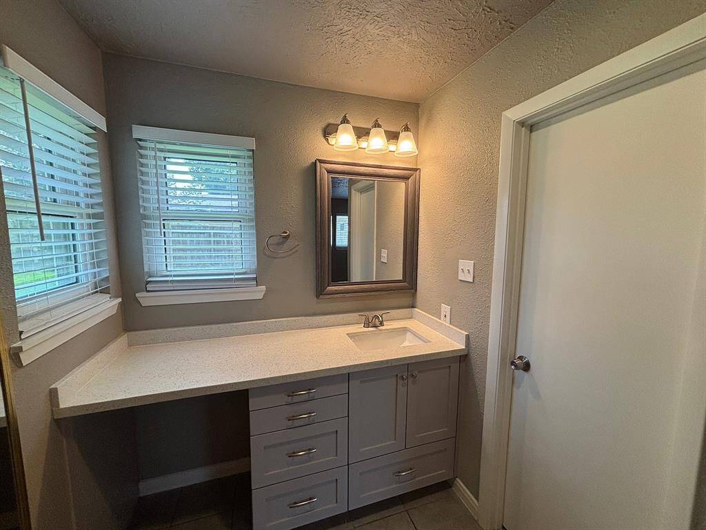 This bathroom features a modern vanity with ample counter space and storage. It includes a sleek sink, a framed mirror, and stylish lighting. Two windows provide natural light, enhancing the room’s bright and airy feel.