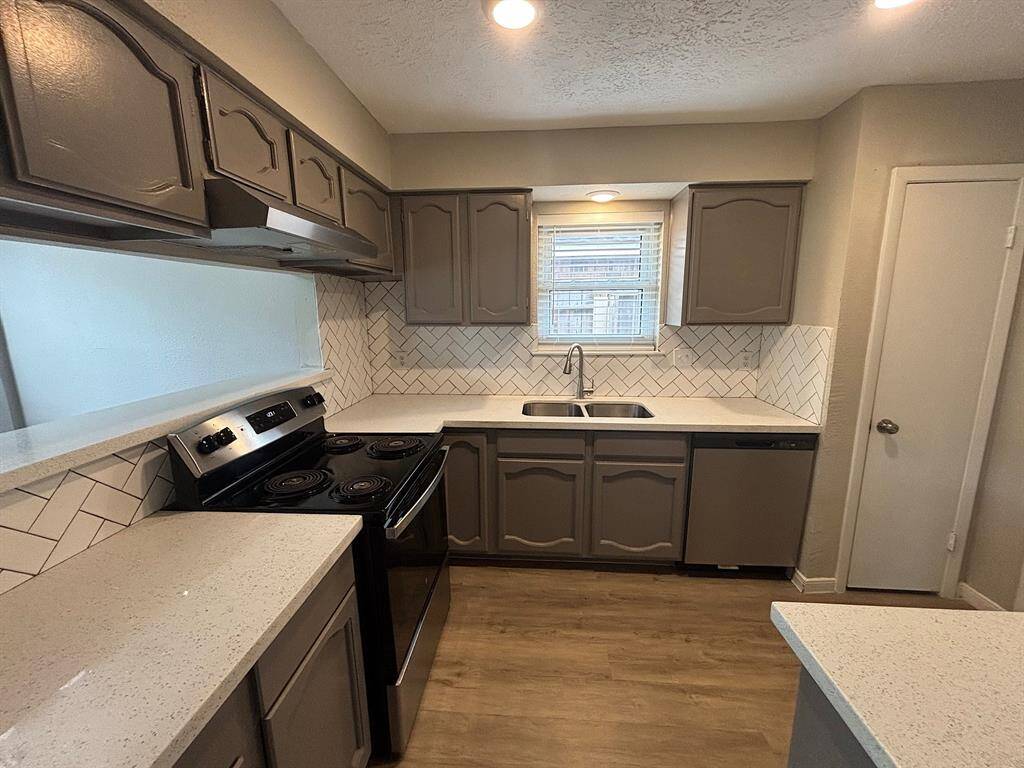 This kitchen features modern gray cabinetry, a sleek stove, and a double sink. The quartz countertops are a light, polished surface complemented by a herringbone tile backsplash. Natural light filters in through a window above the sink, brightening the space.