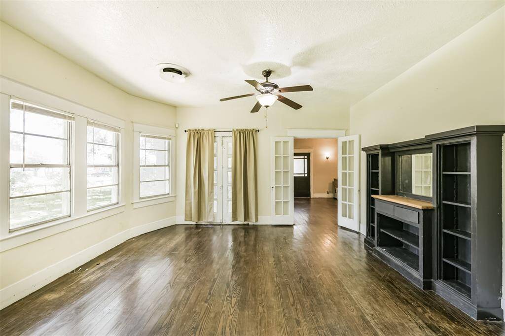 This room features hardwood floors, ample natural light from multiple windows, and a ceiling fan. French doors open to an adjacent space, and there’s a built-in shelving unit with a mantel. Ideal for a cozy dining room.