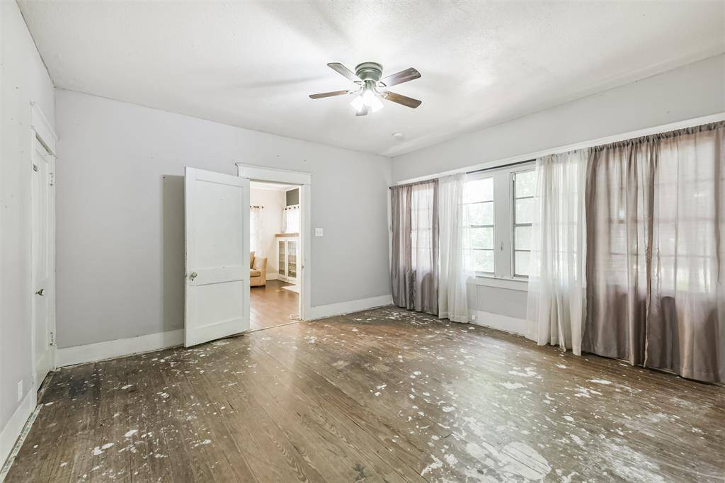 This room is currently the primary bedroom and features large windows with sheer curtains, allowing for plenty of natural light. It has a ceiling fan and hardwood floors that could be refinished. The space connects to the living room through an open door, offering potential for customization.
