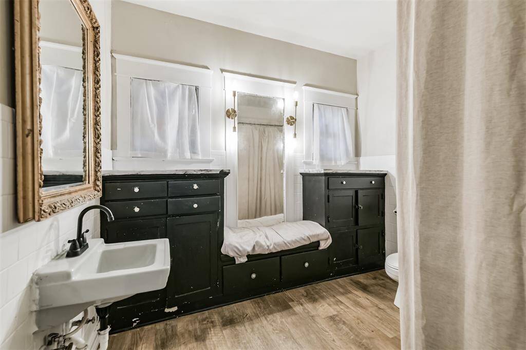 Charming bathroom with vintage appeal, featuring dark wood cabinetry, a classic wall-mounted sink, and a beautiful mirror. Light curtains and wood-look flooring add warmth and character.