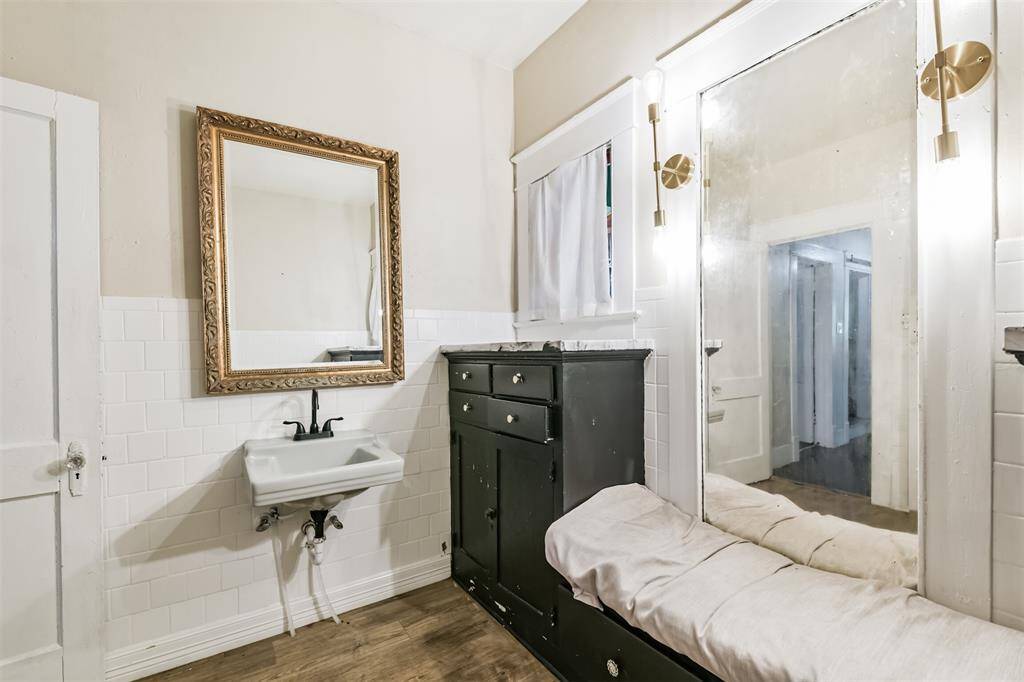 This room features a vintage-style bathroom with a wall-mounted sink, ornate framed mirror, and a black cabinet with marble top. The space includes a large mirror with built-in lighting and a cushioned bench, offering a blend of functionality and classic charm.