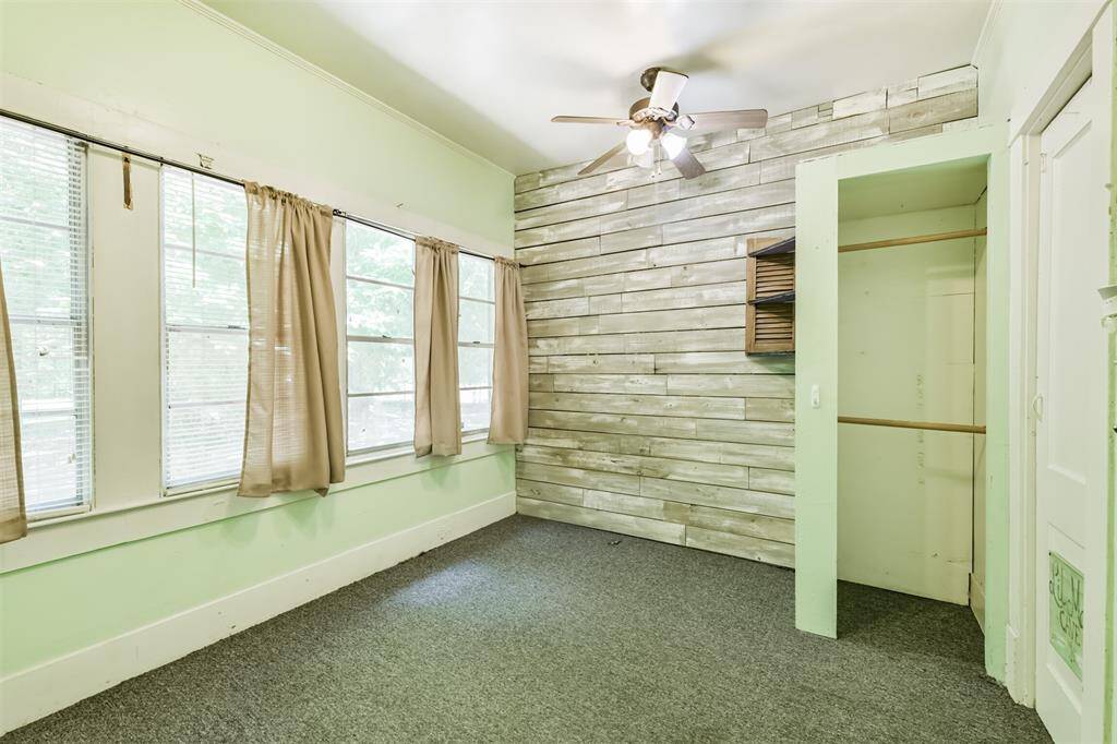 This room features a rustic wood accent wall, large windows with natural light, and neutral carpeting. It includes a ceiling fan and a small open closet space, creating a cozy and functional area.