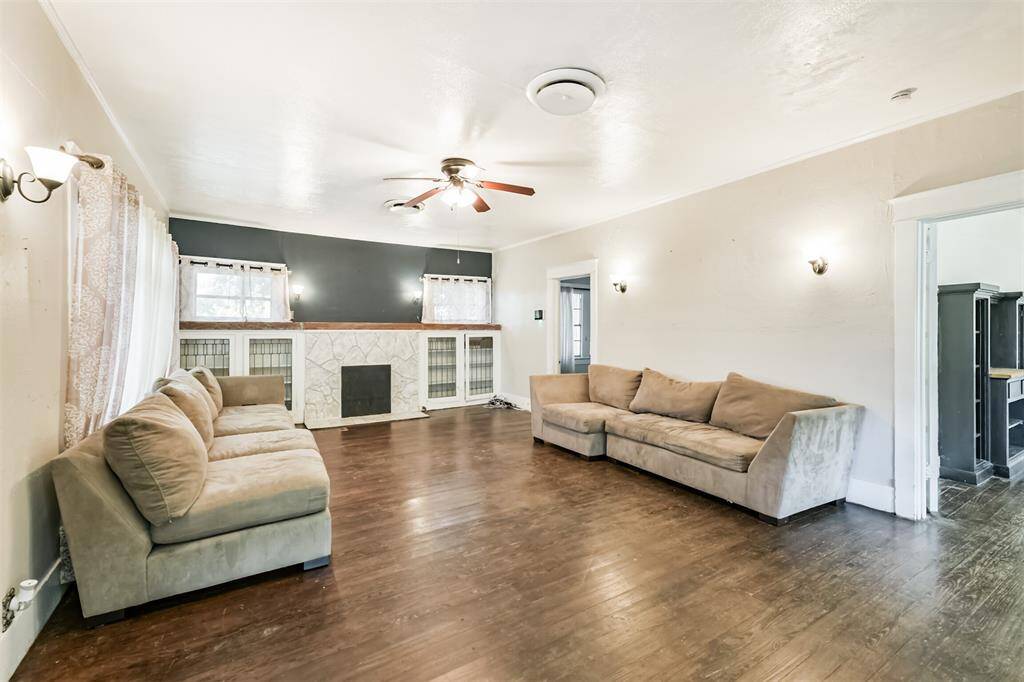 This spacious living room features hardwood floors, a ceiling fan, and a cozy fireplace. It has ample natural light from multiple windows and includes built-in shelving for added storage. The neutral color scheme creates a warm and inviting atmosphere.