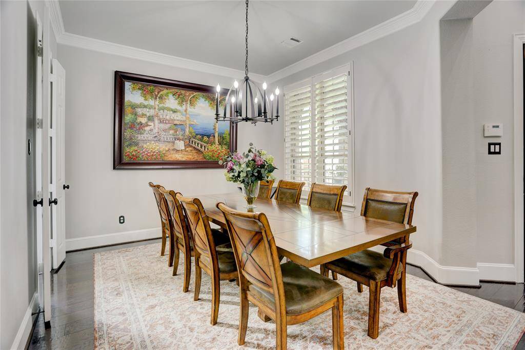 The elegant dining room boasts a large table for eight, soft gray walls, and dark wood flooring. Plantation shutters and tasteful artwork create a warm, refined setting for entertaining.