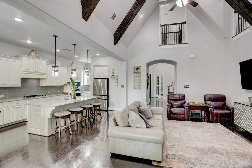 An open-concept layout connects the modern kitchen and cozy living area with leather seating, a mounted TV, and natural light streaming through tall windows. White cabinetry and a spacious island anchor the kitchen.