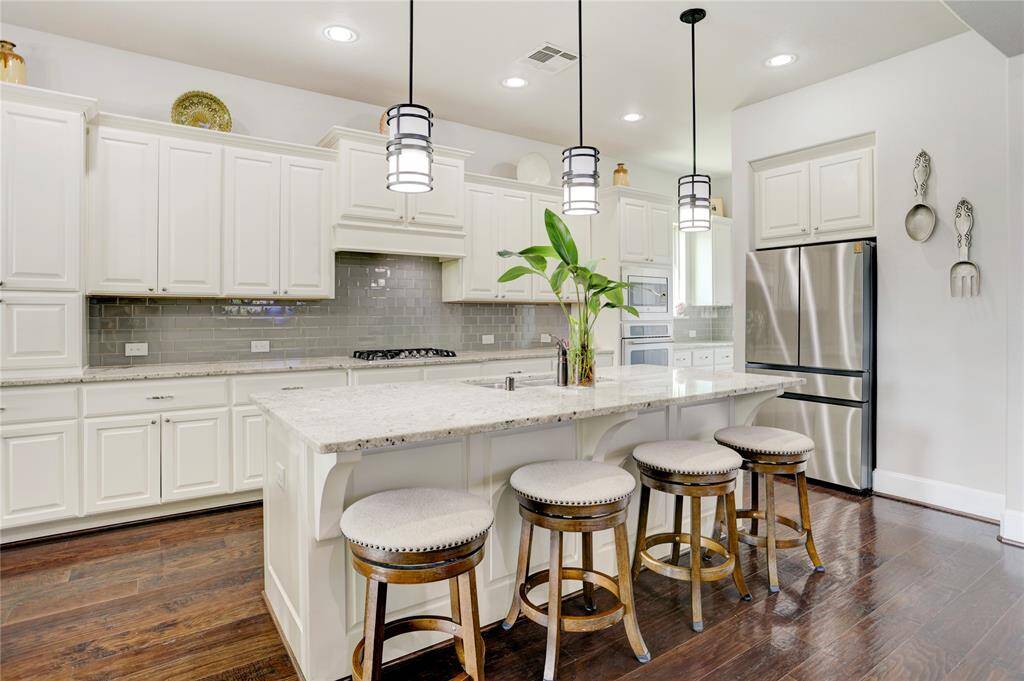 This contemporary kitchen features granite countertops, white cabinetry, and a large island with pendant lighting. Stainless appliances and rich hardwood floors enhance the elegant atmosphere.