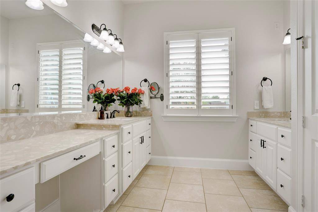 Dual vanities with marble tops, bright lighting, and a large mirror define this elegant bathroom. Tiled floors and a shuttered window add comfort and privacy.