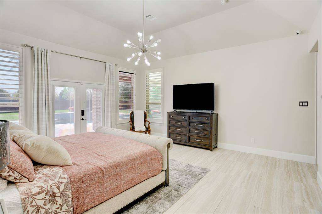 A bright bedroom with soft wood floors, large windows, and double doors leading outdoors. Modern touches include a chandelier, a cozy bed, and sleek furnishings.