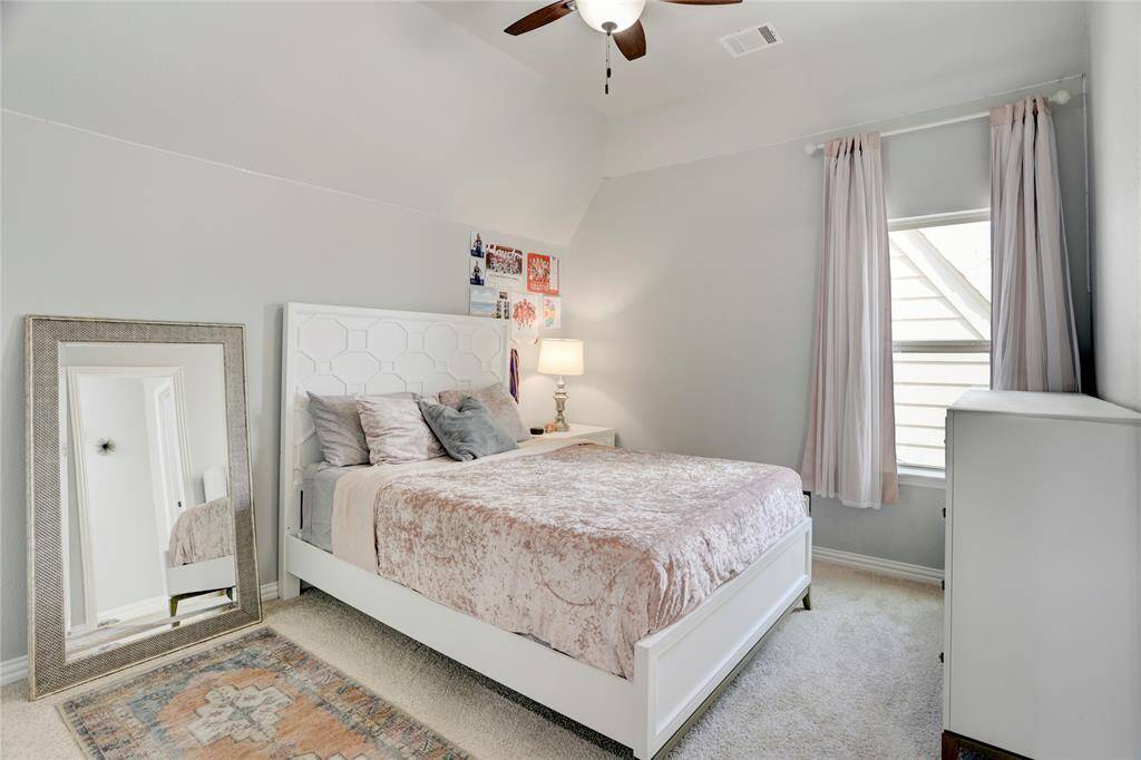 This peaceful bedroom offers a comfy bed, carpeted floors, and a window with light-filtering curtains. A ceiling fan and mirror add functionality and style.