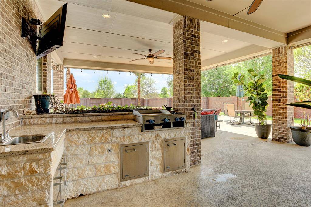 Entertain with ease on this covered patio featuring a full outdoor kitchen, ceiling fans, and generous seating surrounded by greenery and privacy fencing.
