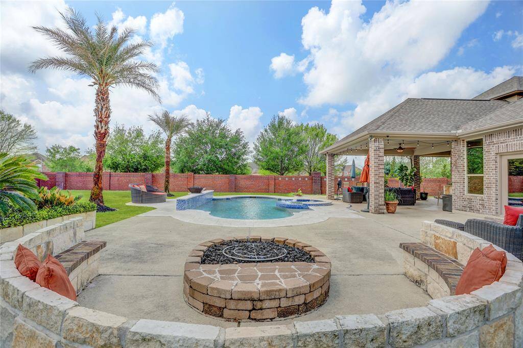 A lush escape with a covered patio, inviting pool and spa, and a built-in fire pit—all bordered by greenery and tall palms for a resort-like ambiance.