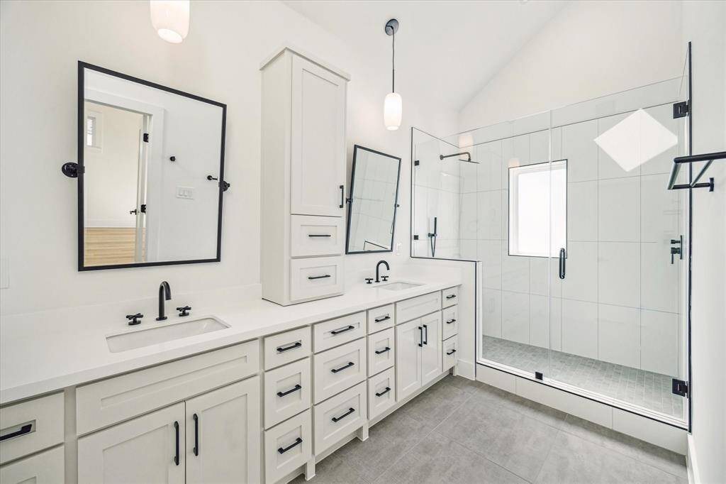 Luxurious primary bath features a vaulted ceiling, sky light, frameless glass walk in shower, dual vanities with coordinating hardware, designer faucets, plumbing accessories, modern light fixtures and separate toilet room