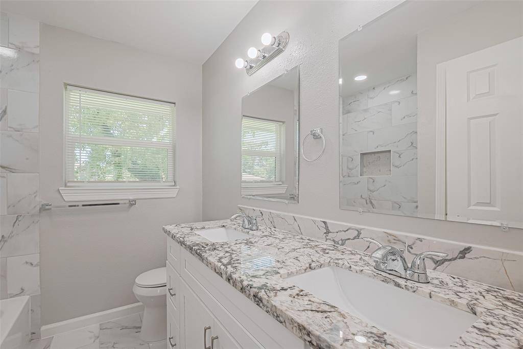 This bathroom also features a dual-sink vanity, twin mirrors, under-cabinet storage, and a window for light and ventilation.