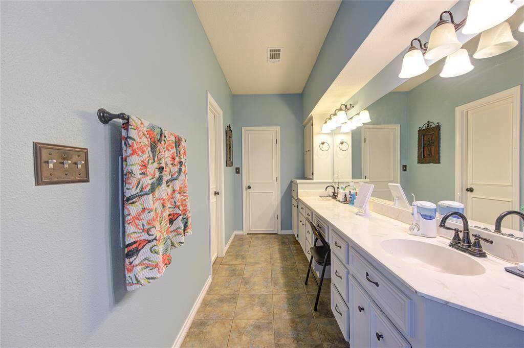 This Primary Bathroom features a long dual-sink vanity with ample storage, elegant lighting fixtures, and a soothing blue color palette. It has tiled flooring and a decorative towel rack, creating a fresh and inviting atmosphere.