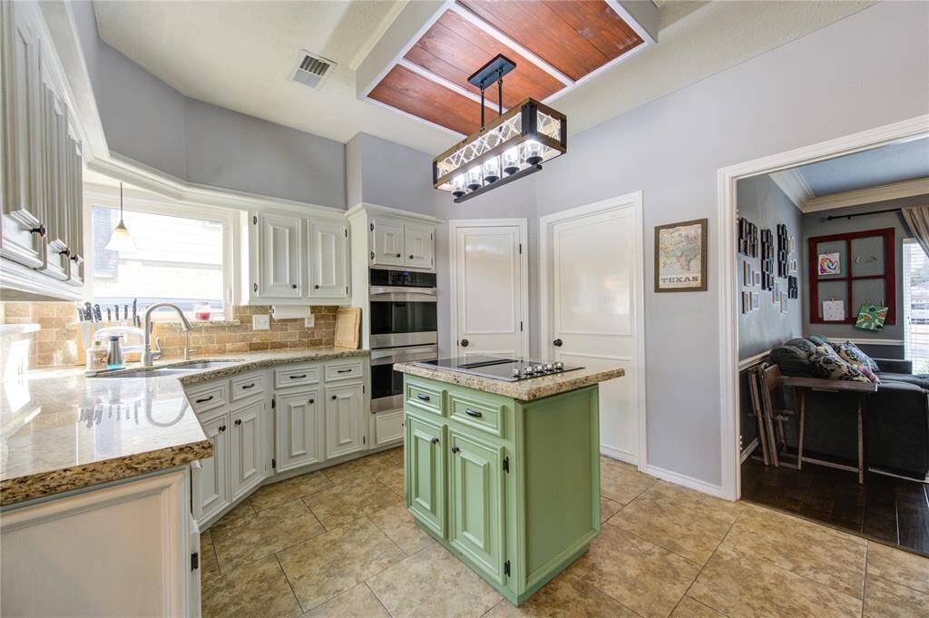 Bright kitchen with white cabinetry and granite countertops, featuring a central green island with a cooktop. The space is enhanced by a unique ceiling design and stylish lighting, opening into a cozy living area. Double stanless steel ovens!