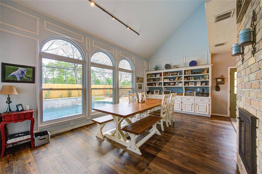 This inviting dining area features a high ceiling and large arched windows that offer a view of the backyard and pool. The space includes ample room for an oversized dining table, built-in shelving for display, and a cozy brick fireplace, all set on warm luxury vinyl plank flooring.
