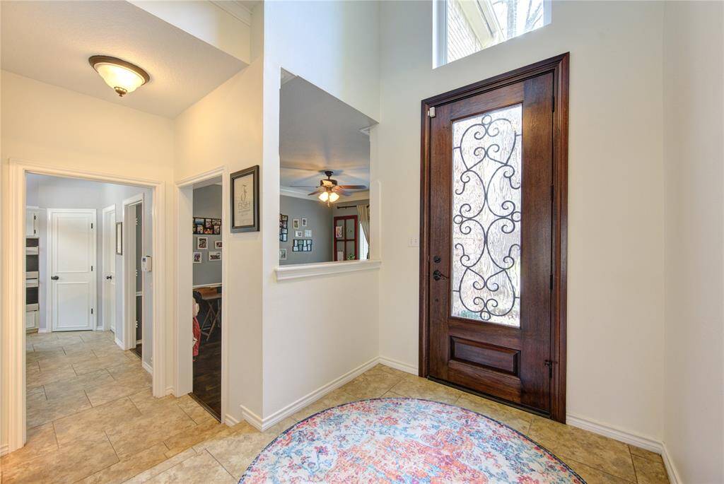 This welcoming entryway features a stylish wooden door with decorative glass, tile flooring, and a cozy, light-filled atmosphere. The space opens into a well-lit interior with a view of the adjoining rooms, offering a glimpse of the home's layout.